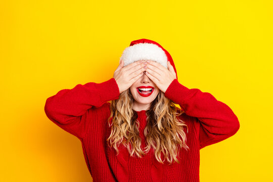 Joyful woman in Santa hat covering her eyes in playful holiday surprise wearing red sweater on bright yellow background