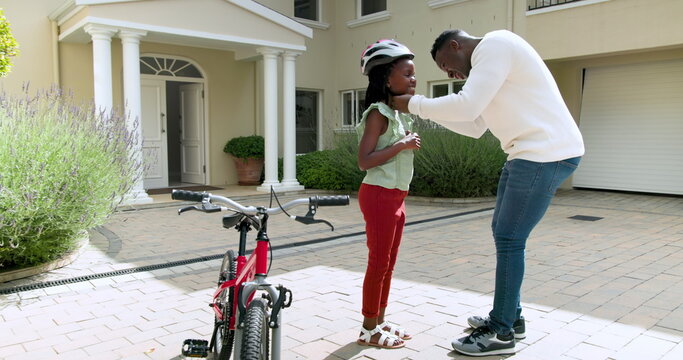 Happy black father teaching his smiling daughter to ride a bike outside their home, girl wearing hel