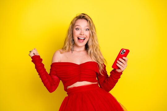 Excited young woman in red dress holding smartphone smiling against a vibrant yellow background