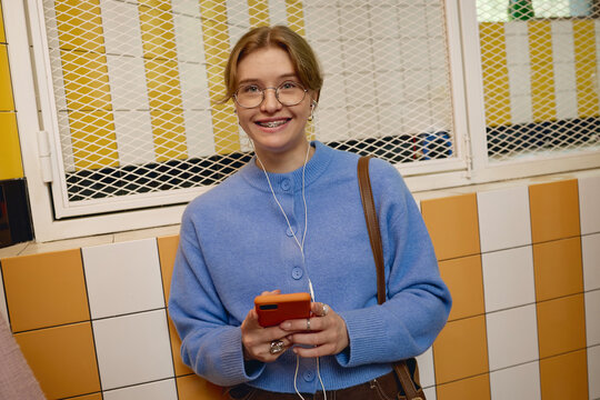 Portrait of smiling Caucasian young adult woman wearing eyeglasses with braces holding smartphone and earphones. Visual representing mobile technology, ideal for digital app advertising