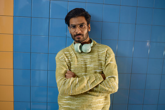 Portrait of young adult South Asian man wearing eyeglasses and headphones around neck, looking at camera with crossed arms. Image suitable for lifestyle, audio tech or music marketing