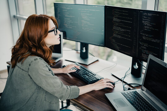 Female software developer coding on a multi-monitor workstation with laptop, focused programmer working at desk in a modern office workspace