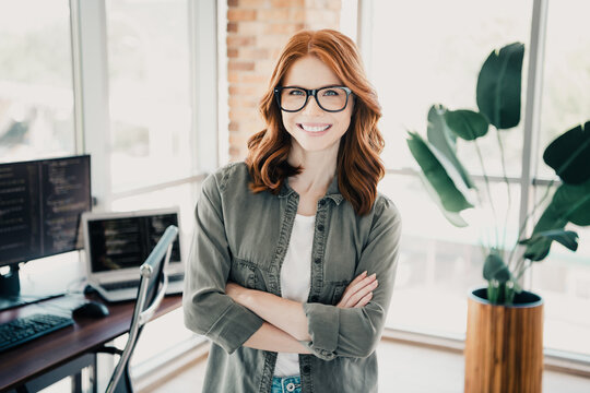 Smiling redhead female developer in a modern office with laptop and monitors, standing confidently with arms crossed wearing glasses