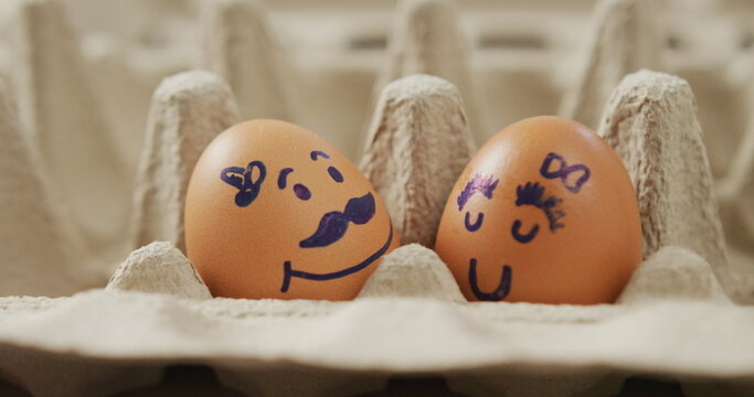 Image of close up of two brown eggs with drawn faces in egg carton background