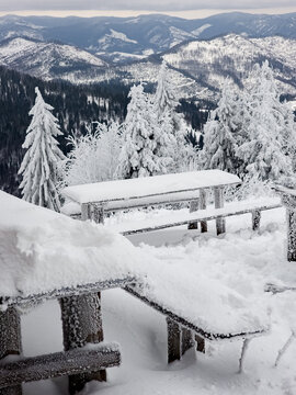 Snow-covered wooden picnic tables and benches on a mountain viewpoint during winter