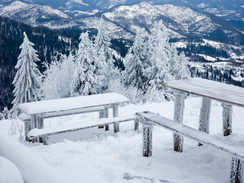 Snow-covered wooden picnic tables and benches on a mountain viewpoint during winter