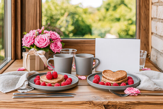 A romantic breakfast scene featuring two plates of heart shaped pancakes topped with raspberries mugs of coffee a bouquet of pink roses and a blank greeting card on a rustic wooden windowsill