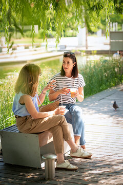 Two female friends relaxing in a public park, having a conversation and drinking iced drinks on a sunny day