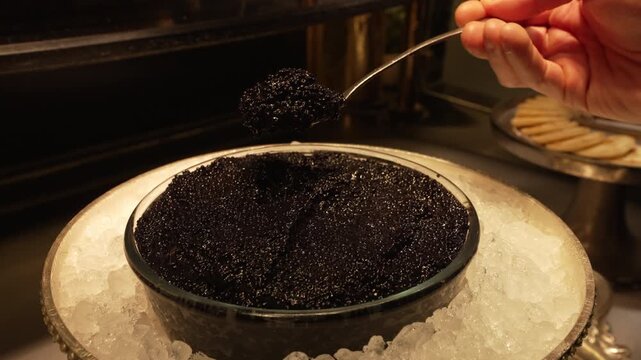Bowl of black caviar on ice with spoon lifting a portion and blinis nearby