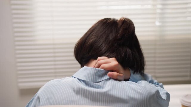 Back view of Asian woman holding her stiff neck in pain while sitting at office desk, experiencing severe muscle strain and fatigue.