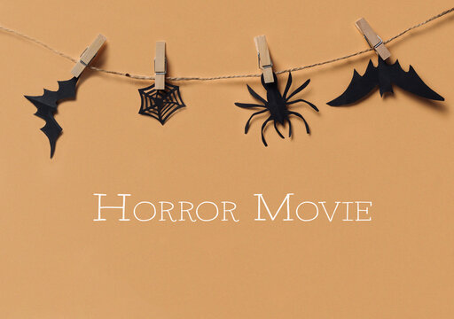 Paper cutouts of bats, a spider, and a web hang on a string with clothespins against a beige background, suggesting Halloween decor.