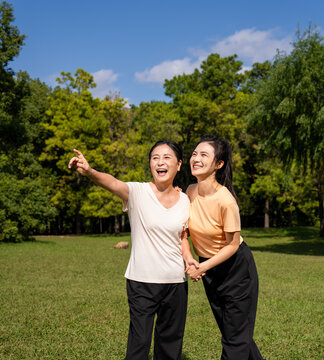  Happy Asian senior mother and adult daughter holding hands, smiling together in green park on sunny day, family bonding outdoor portrait