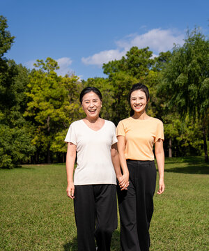  Happy Asian senior mother and adult daughter holding hands, smiling together in green park on sunny day, family bonding outdoor portrait