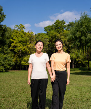  Happy Asian senior mother and adult daughter holding hands, smiling together in green park on sunny day, family bonding outdoor portrait