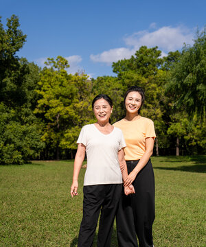  Happy Asian senior mother and adult daughter holding hands, smiling together in green park on sunny day, family bonding outdoor portrait