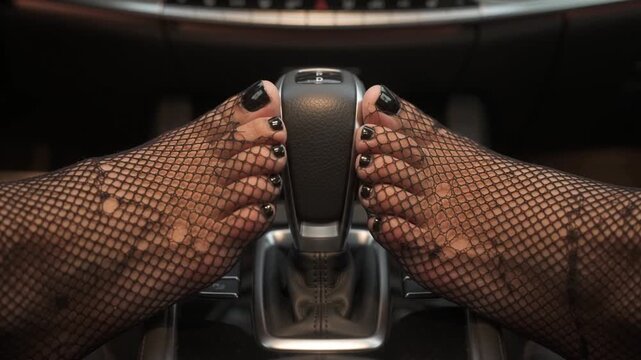Commercial stock video of two feet wearing fine black fishnet stockings and black-polished nails pressing inward on a luxury car gear shift knob.