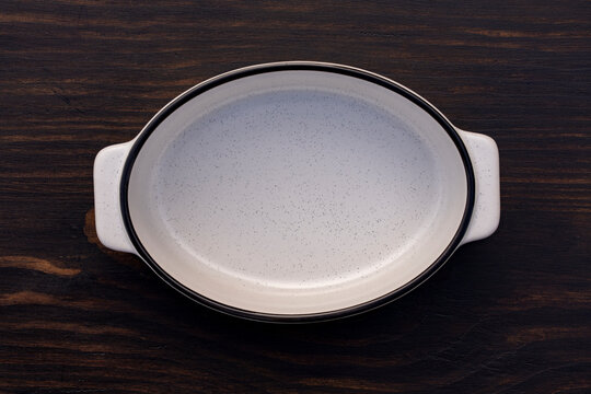 Empty white ceramic baking dish on wooden background top view, closeup. Empty oval ceramic casserole dish with speckles and handles placed on dark wooden table, flat lay copy space