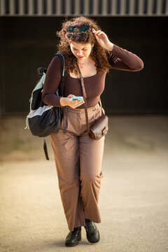 Young woman walking outdoors while using smartphone and carrying backpack