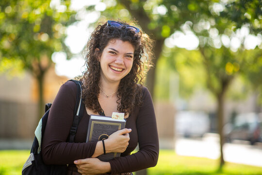 Cheerful young female student laughing with book and backpack outdoors on campus