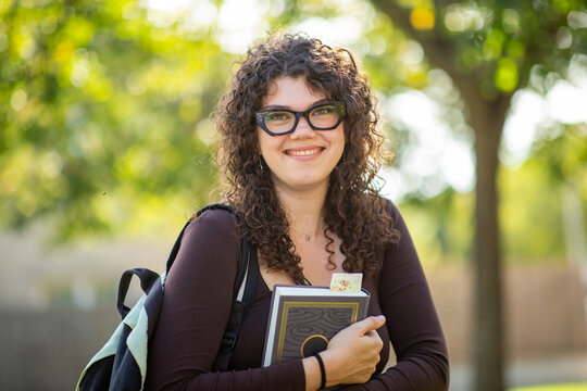 Smiling young female student with glasses holding a book on sunny university campus