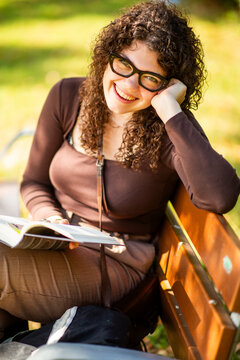 Cheerful young woman with glasses relaxing with book on park bench