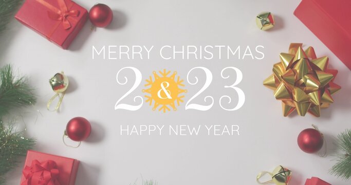 Red gift boxes and gold bells framing Merry Christmas 2023 lettering on pale surface, copy space