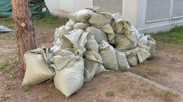 Pile of filled grey polypropylene sandbags stacks on ground near tree trunk and industrial building outdoors. Construction material supply, flood protection barrier or utility engineering debris.