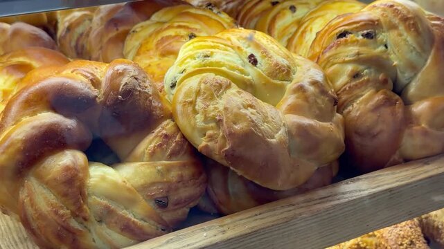 Freshly baked golden braided buns with raisins lie inside a wooden tray behind glass at a local bakery store. Delicious sweet traditional pastry confectionery retail display counter layout.