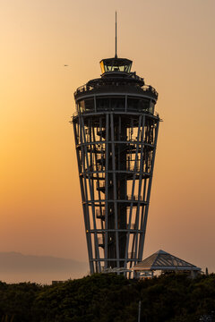 Steel lattice observation tower and lighthouse silhouetted against warm golden sunset sky, visitors on upper deck overlooking foliage below