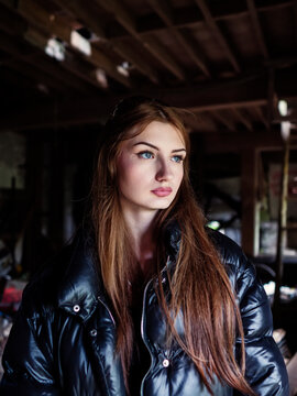 Young woman with long brown hair wearing black puffer jacket. She stands in dimly lit, cluttered abandoned building. Her eyes are closed, expression serene and thoughtful. Scene evokes mystery