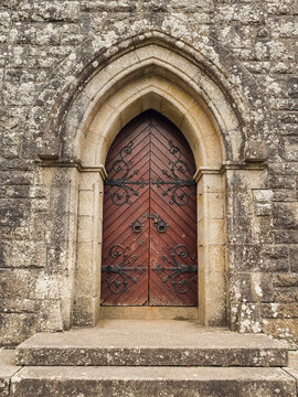 Old stone church with arched wooden door. Steps lead up to entrance. Moss grows on stones. Trees stand in background. Gravel surrounds foundation