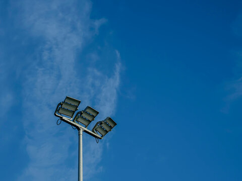 Tall floodlight stands against bright blue sky with wispy clouds. Metal pole supports multiple lamps pointing upward. in the scene. Ideal for sports, stadium, or event-related projects