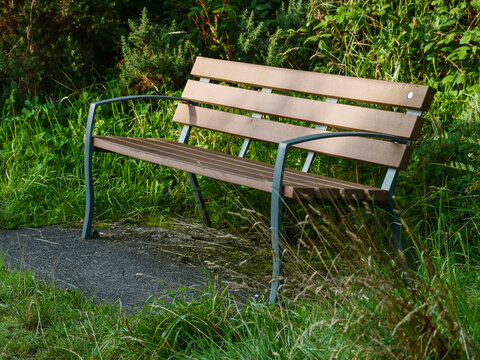 Wooden bench with metal arms sits beside gravel path. Green grass and bushes surround the bench. It offers quiet solitude for peaceful moments in nature. Perfect for parks, gardens
