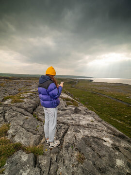 Person stands on a rocky cliff, gazing at a vast coastal landscape. The sky is overcast, with clouds hanging low above the sea. The individual wears a bright colorful jacket and a yellow beanie