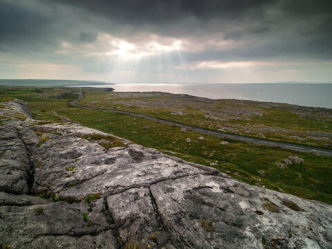 Sunlight breaks through stormy clouds over rugged coastal cliffs. Winding road cuts across green hills toward the ocean. Weathered stone textures dominate the foreground