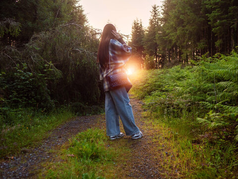 Silhouette of woman standing on dirt path, sunlight filters through trees
