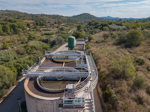 Circular clarifier tanks and an activated sludge aeration basin at a wastewater treatment plant surrounded by green hills and shrubland under a blue sky