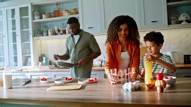 African american family breakfast kitchen. Dad serving eggs, boy pouring juice.