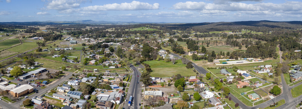 Aerial drone panorama of Beaufort in Victoria, Australia, the regional railway line parallel to Western Highway running through the town centre, surrounding rolling pastoral hills and native forest