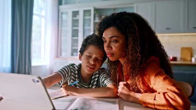 Single mom tutoring son homework tablet kitchen. Boy laughing homeschool lesson.