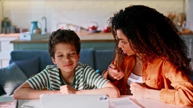 Boy writing homework tablet kitchen table. Mom explaining lesson smiling son.