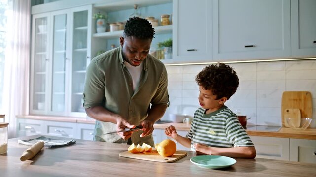 Dad teaching boy cutting orange knife kitchen. African american family cooking.