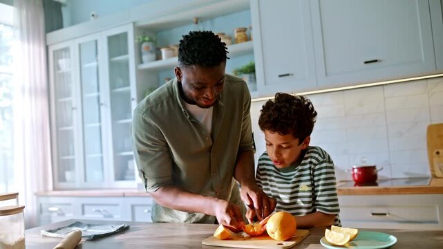 Son cutting orange knife dad help. Happy family cooking together home kitchen.
