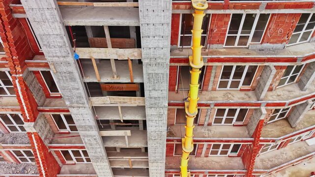 A yellow debris chute runs down the facade of a brick apartment building, beside unfinished concrete balconies and rows of windows during daytime construction.