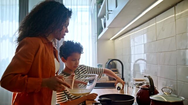 Mother pouring whisked eggs frying pan. Son squeezes oil, cooking omelette home.