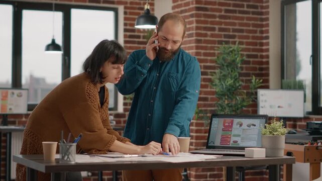 Phone, communication and teamwork as a man speaks on a smartphone while a woman reviews charts and takes notes in an office. Business coordination, multitasking and project planning during a workday.