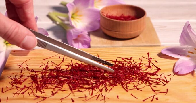 Woman taking saffron with tweezers near crocus flowers at light wooden table, closeup