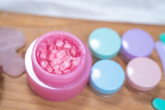 Close up of thick pink gel with beads filling a small open jar on a wooden surface, with pastel lidded containers nearby, a concept of texture marketing for indie cosmetic brands.