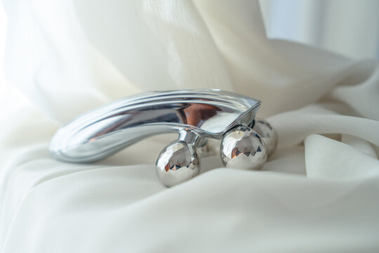 A silver handheld massage roller with faceted metal balls rests on draped cream silk fabric, a beauty tool for skincare, lymphatic drainage and spa self care at home.