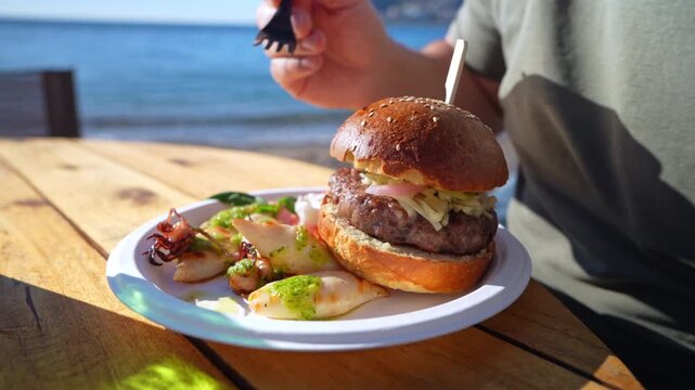 A man picks up a piece of grilled squid with a fork from a paper plate holding a sesame bun beef burger and grilled squid, at an outdoor table beside the blue sea.